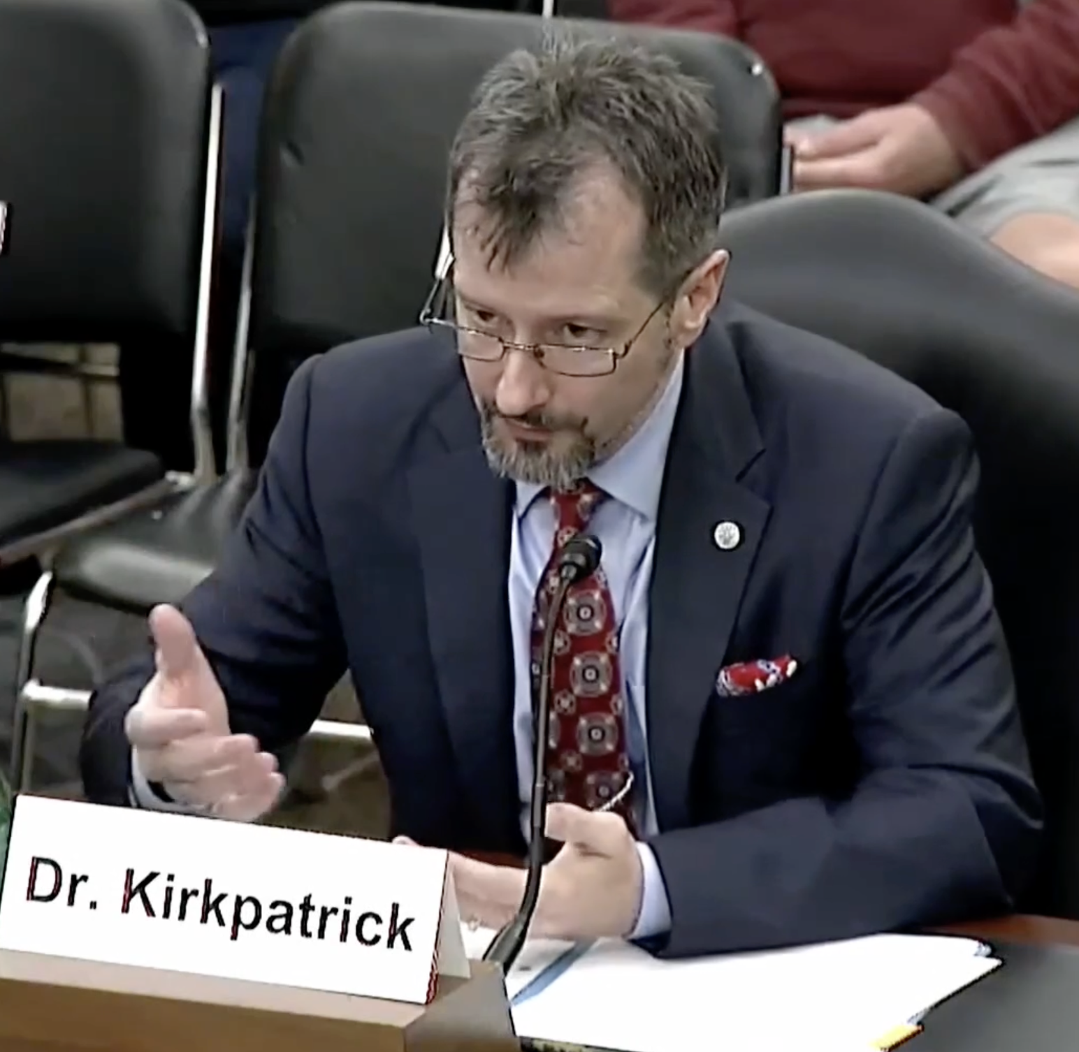 Dr. Sean Kirkpatrick sitting at a desk with a microphone. It appears he is speaking in front of Congress.