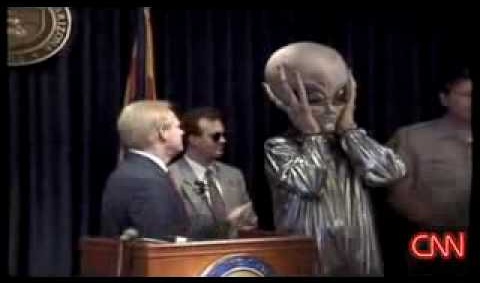 A politician stands in front of a podium for a press event as a person stands beside him in an exaggerated alien costume. This is a historical event that actually happened in Arizona after the Pheonix Lights mass sighting.